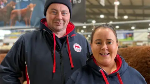 A man and woman in branded navy and red hoodies stand inside a livestock hall in front of cattle, with a banner reading “Lisnamul Farmers” visible in the background.