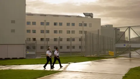 Two prison officers, walking past a prison, with a block, with windows and a fence to the right. The ground is wet. 