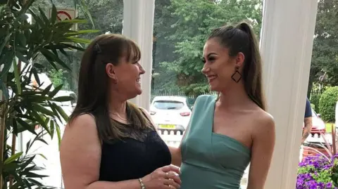 Family handout Maria Britton on the left, wearing a black dress with Emily on the right, wearing a teal colour cocktail style dress. They are in a home in front of a window.