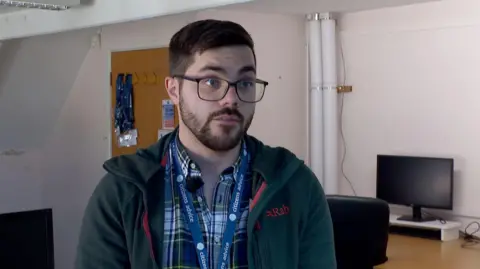 A man with short dark brown hair wearing rectangular glasses, a plaid shirt underneath a green hoodie standing in an office room.