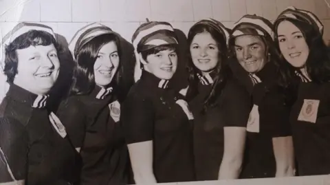 Anne Kane Mary and her synchronised swimming team standing in a row smiling at the camera. They are wearing dark coloured uniforms and stripy hats. 