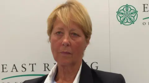 Conservative councillor Victoria Aitken - a woman with short blonde hair wearing a dark blazer and a striped shirt. She is sitting in front of a board with East Riding of Yorkshire Council's logo on it. 