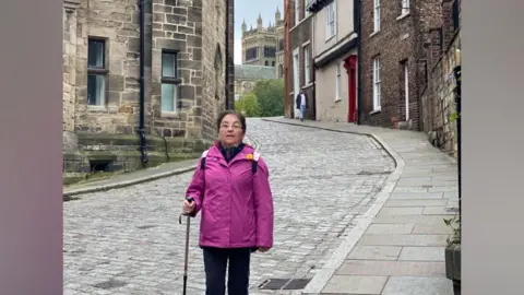 Supplied Doreen Park has short brown hair and glasses. She is wearing a pink jacket and black trousers as she poses for a photo down Owengate. The street is cobbled and surrounded by stone and brick houses. Part of Durham Cathedral is visible at the top of the street.