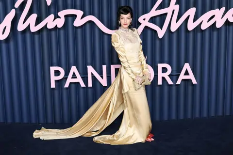 Getty Lily Allen wearing a gold dress with red shoes and a matching gold clutch, with her dark hair curled and tied back