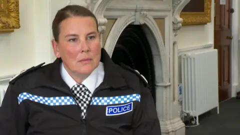 A woman with tied-back dark hair, wearing a police uniform, is standing in a room near what looks like an old-fashioned fireplace. She is looking past the camera at something.