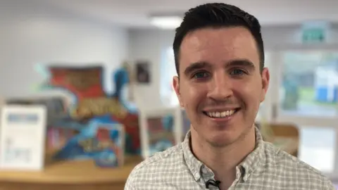 A fresh-faced teacher with short dark hair smiling to the camera. He is standing in a school classroom with children's reading books in the background and wearing an open collar cream-patterned shirt