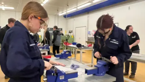 Emma Baugh/BBC In a workshop two female apprenticeships wearing dark blue overalls are working with bench vices, in the room there are several benches