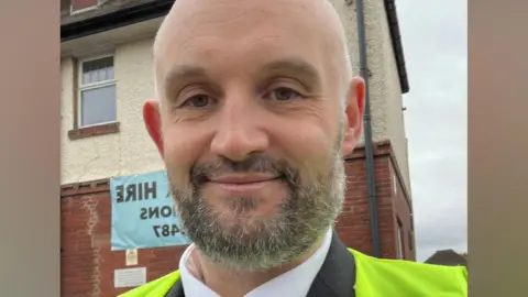 Mark Barber A bald man with a dark grey beard takes a selfie while wearing a yellow high viz jacket.