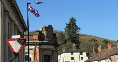 A skyline of Church Stretton can be seen, with a big green hill behind. It also shows buildings in the foreground, with old stonework and white and red brick buildings. There is also a flag of the United Kingdom tied up to the top of a black lamppost, and a red 'Give Way' traffic sign.
