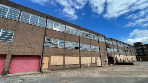 A large brick building with long rows of windows. The wide concrete yard in front is empty. The image has been taken on a sunny day with a blue sky overhead.