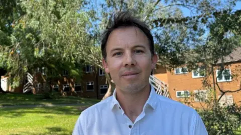 A man with short black hair wearing a white shirt. He is stood in a park looking into the camera with a row of houses behind him