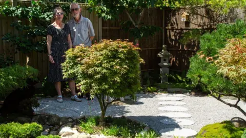 Roger Harris An elderly man and wife stand at the back of a garden where small ornamental trees and grasses grow. There is bamboo fencing, a stone lantern, and round paving stones in a row on top of gravel. 