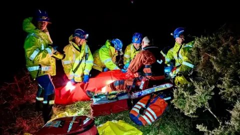 Kingsbridge Coastguard Rescue The picture shows a group of Coastguard rescue crews working around a casualty in the dark, lit only by their powerful head‑torches and the glow from their gear. The rescuers are wearing bright yellow jackets, helmets and protective trousers, and their reflective strips shine brightly against the black night behind them.