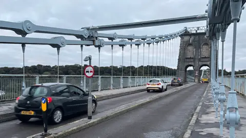 Three cars cross Menai Bridge with its huge blue grey metal hanger clearly visible, holding the road in place
