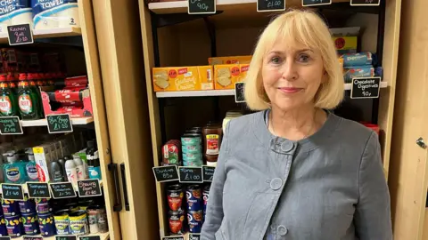 A woman wearing a grey blazer with a blonde bob smiling. She is standing in front of shelves in a shop. The shelves have a range of tins and packets on them including for biscuits and washing up liquid.