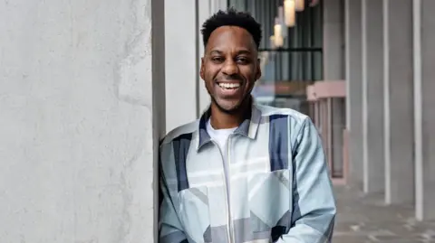 Elliott Rae Elliott Rae is standing outside in an urban setting, smiling. He has black hair and is leaning casually against a concrete column. He is wearing a light-coloured jacket with a geometric pattern in shades of blue, grey, and white, over a white shirt.