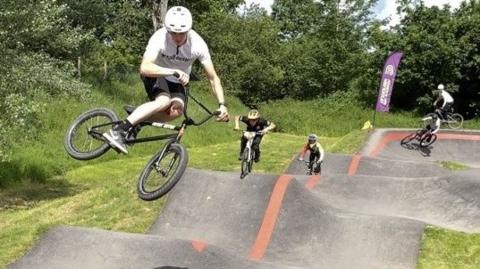 BMX riders riding the rolling hills of the pump track with one jumping high in the end and kicking out their back tyre towards the camera