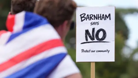 PA Media A protester in Barnham wears a Union Flag as a cape and looks towards a sign that says "Barnham says no. Protect our community".