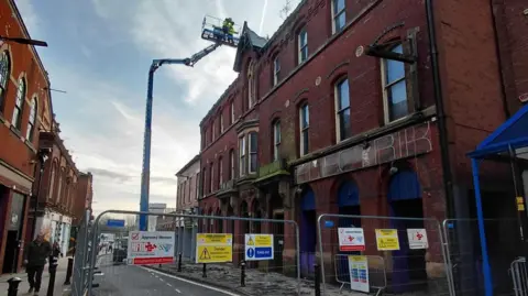 Wigan Council Facebook A tall red brick building can be seen but in front are metal fences with warning signs all over reading 'danger'. A crane can be seen as workers in hi-vis jackets.