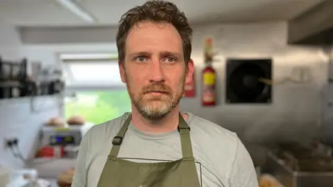 BBC/Seb Cheer A man with brown hair and a beard looks into the camera. He is standing in a small commercial kitchen, wearing a grey T-shirt and green apron.