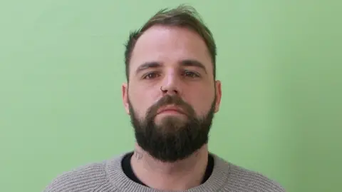 Guernsey Police Jamie Ferbrache in a police custody photograph. He is standing in front of a green background. He is wearing a beige jumper. He has short brown hair and a beard. He has a nose piercing and a neck tattoo.