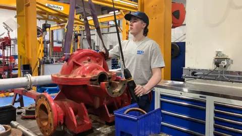The photo is taking in a factory. A man is working at a large red pump which he is about to lift with a crane system that he is controlling using a button. You can see work tools and yellow steel pillars in the background. The man is wearing a black baseball cap and has long brown hair.