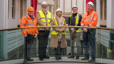 STACK Councillor Nigel Stansfield, Councillor James Petter, Gemma Dishman, Paul Wright, Stuart Timmiss, all standing on a glass walkway, wearing hard hats, smiling and looking at the camera. 