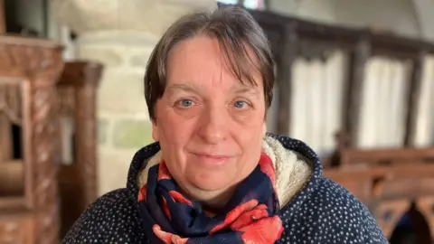 Sara Burrows pictured at the church. She is wearing a blue jumper with white dots and a fleece like hood. She is wearing a scarf with a poppy design on it.