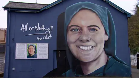 A large wall mural of Sr Clare in the Brandywell in Derry. A painted portrait of St Clare smiling is on one side of the wall with 'All or Nothing' is written in spray paint beside her.