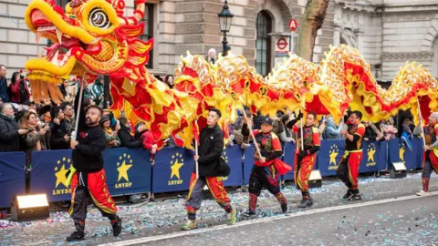 London Parade Peter Wai, a man with short dark hair and beard, walks at the front of a red and yellow Chinese style dragon. He is holding a pole attached to the head and dressed in red and black clothes