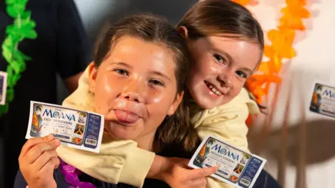 A close-up image of two girls holding 'Moana Exclusive Screening' cinema tickets. One is poking her tongue out. They are both smiling and looking into the camera.