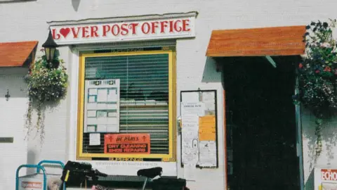 Nick Gibbs Lover's old Post Office, with a red sign reading "Lover Post Office" with a heart in place of the first "O". There are flower baskets on the white walls.
