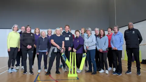 A group of walking cricket players stand in a group holding bright yellow plastic cricket bats and yellow cricket stumps in an indoor sports hall