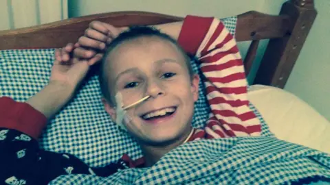 Robbie is grinning and looking at the camera, but this time he is lying in a wood-frame single bed. His hair is shorter and he has a tube taped to the side of his face, feeding into his right nostril. Robbie has his arms raised above his head in a relaxed position and wears a long-sleeved pyjama top, one sleeve is red and white stripes, the other is black with a white skull print. His head is resting on a blue and white gingham print pillow and he is under a matching blue and white gingham duvet cover.