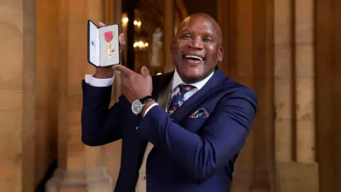 PA Media Devon Malcom smiles widely at the camera and is dressed in a navy suit with a matching patterned navy tie and handkerchief in his pocket. He is holding up his gold medal.