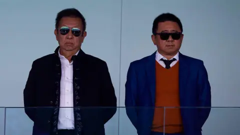 Getty Images Two men, wearing sunglasses, look ahead behind a glass barrier at a football stadium.