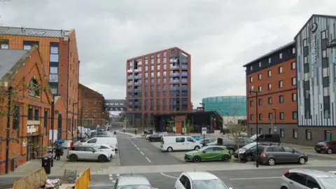 Gloucester City Council/Rokeby Merchant Developments The view from St Anne Way towards Gloucester Docks. It shows a large car park in the centre, with Downing's Malthouse on the left and a Premier Inn hotel on the right. In the distance there is a tall tower block with lots of glass windows.