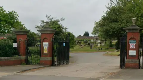 The entrance to Thorntree Cemetery in Middlesbrough features three red-brick columns topped with ornate stone work and with tall, black metal gates between them. The gates to the site are open and a roadway leads to dozens of gravestones dotted around a green field beyond.