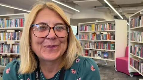 Andrew Turner/BBC Kerry Murray a lady sitting in a library who is smiling and looking at the camera. She is wearing a teal floral top and has shoulder length blonde hair.