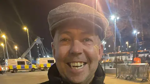 Plymouth Argyle fan Steve smiles as he leaves the Etihad Stadium in Manchester. He is wearing a grey flat cap. Two police vans are behind him and a safety steward is walking past some fencing over Steve's right shoulder. It is night time and several street lights are on.