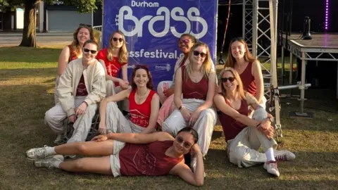 The nine female members of Brazen Brass Band smile as they sit in front of a purple banner reading Durham Brass Festival. One member of the band is lying on her side in front of the others, with her head resting on her left hand.
