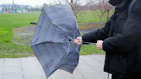 A multi-coloured umbrella is bent in the wind. A person in a black coat is holding it. They are outside, the background is a grass area with bare trees. 