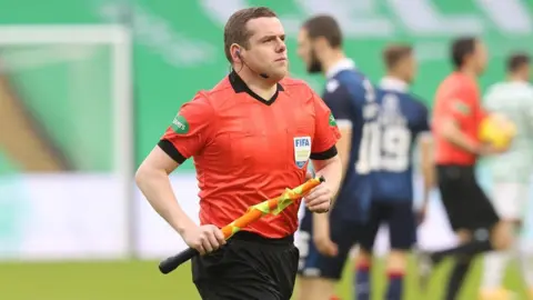 PA Media A man with dark hair jogs across a football pitch while holding a fluorescent referee's flag and red and black refereeing uniform. Footballer players, a stand and goal posts can be seen in the background. 