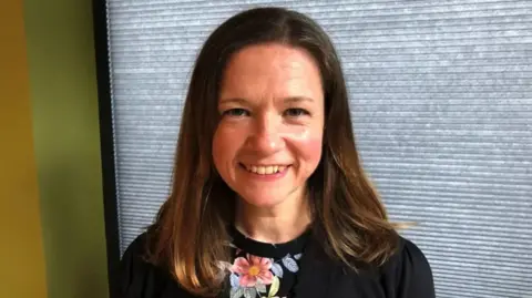 Philippa Cotterill is looking at the camera. She is smiling with her brown shoulder-length hair framing her face. She has a black top and in the middle it has pink flowers with blue and green leaves. She is stood in front of black blinds that are closed with a green wall to the left of her.