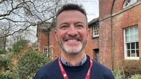 ELLEN KNIGHT/BBC Derw pictured looking into the camera and smiling. He's wearing a navy knitted jumper with a blue and white striped collared shirt on underneath. He's also wearing a red lanyard with 'Attingham Park' written on it. He's stood in front of an old, red brick cottage, which has white framed windows. There is a tree on the left side of the photo, with its branches reaching across the top of the image. The sky is grey and overcast. 