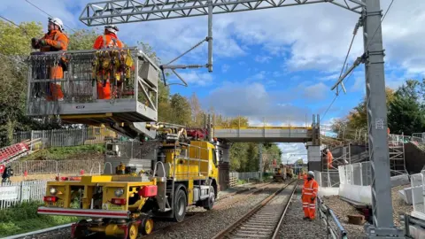 Network Rail A yellow lorry on a rail track carrying people involved in upgrade work on overhead cables. They wear orange boiler suits and hard hats.