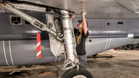 Lightning Association Grace wearing a hat, goggles, a black t-shirt and jeans with her hands against the bottom of a wing of a jet. She is looking up and using a tool. 