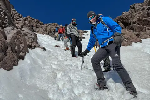The Content Creators The photo is taken on the side of a snowy mountain, with some jagged rocks on show, and a deep blue cloudless sky. The photo has been taken from below with Gerwyn stood in the foreground of the photo, and five other men visible to varying degrees in the background. Gerwyn is leaning on an ice axe which has been wedged in the snow. Every man is wearing warm winter gear and sunglasses. 
