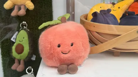 The very round and orange Jellycat fuzzy peach toy sits smiling in a shop window display with other Jellycat toys including an avocado and a smiling banana