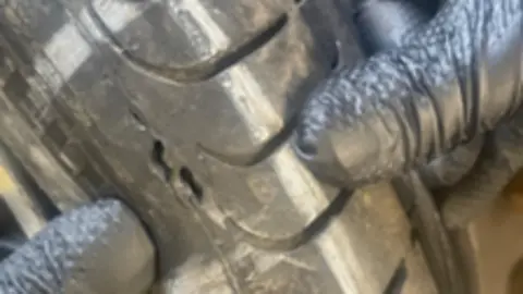 John Smyth A close up of a punctured tyre. Gloved hands are touching the tyre. 